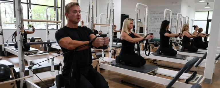 People exercising on Pilates reformer machines in a bright studio, all wearing black workout clothes.