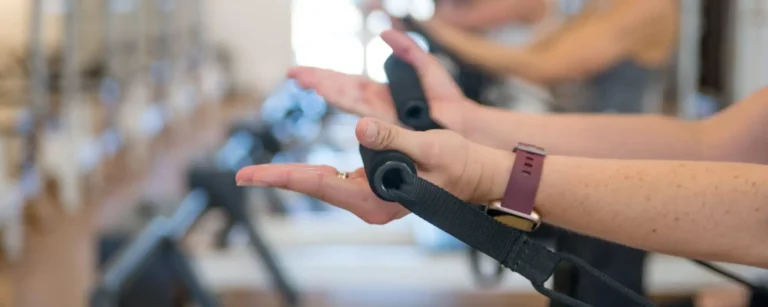 Close-up of hands gripping Pilates reformer straps during a workout, with blurred gym equipment in the background.