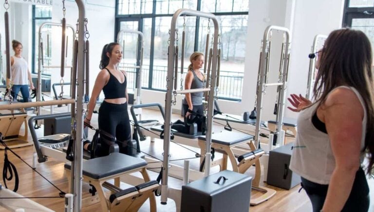 Women in workout clothes use Pilates reformer machines while an instructor leads a class in a bright studio.