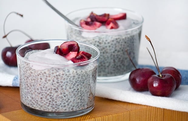 Two glass cups of chia pudding topped with cherries, with fresh cherries beside them on a wooden surface.