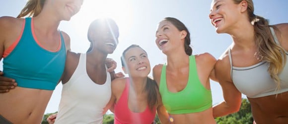 Five women in sports bras smiling and standing together outdoors with sunlight in the background.