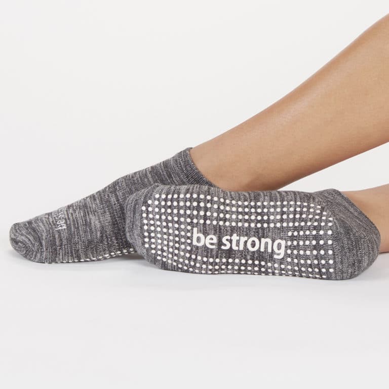 Gray grip socks on feet, with “be strong” written on the sole in white text, against a white background.