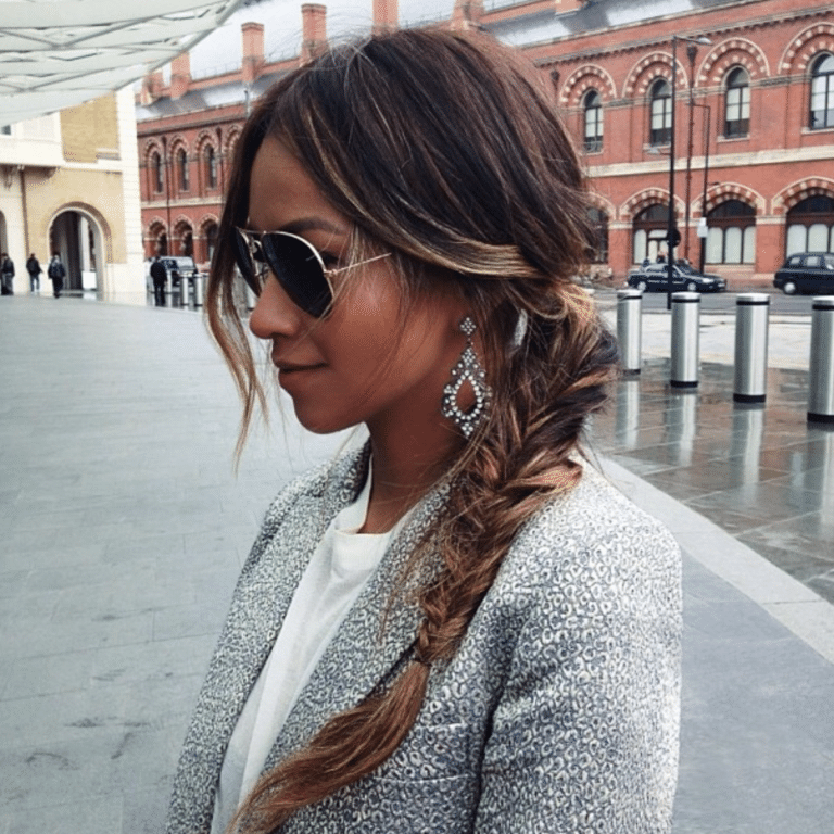 Woman with a fishtail braid, sunglasses, and earrings, wearing a patterned blazer, standing outdoors by brick buildings.