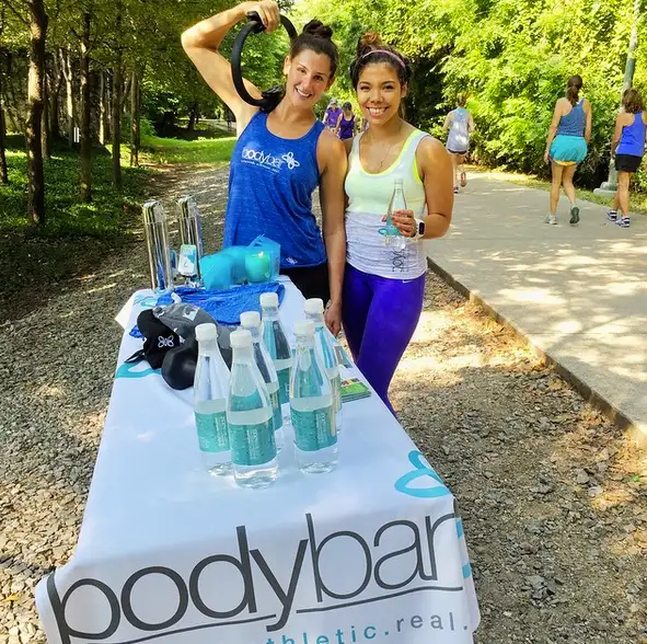 Two women stand by a table with water bottles and fitness gear outdoors, smiling on a sunny path surrounded by trees.