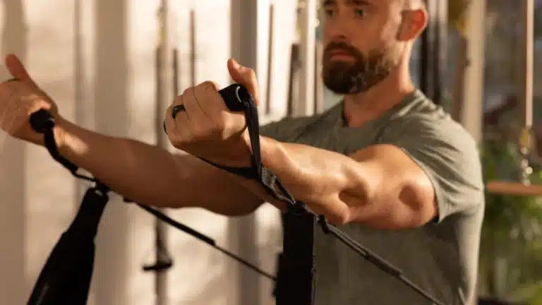 A man with a beard uses resistance bands for exercise, extending his arms forward in a well-lit room.