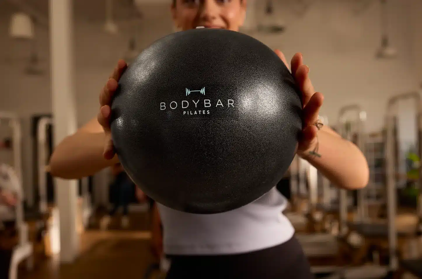 Person holding a black BODYBAR Pilates exercise ball in a gym, with pilates equipment visible in the background.