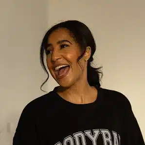 A woman with dark hair in a ponytail smiles widely, wearing a black shirt with white text, against a plain background.