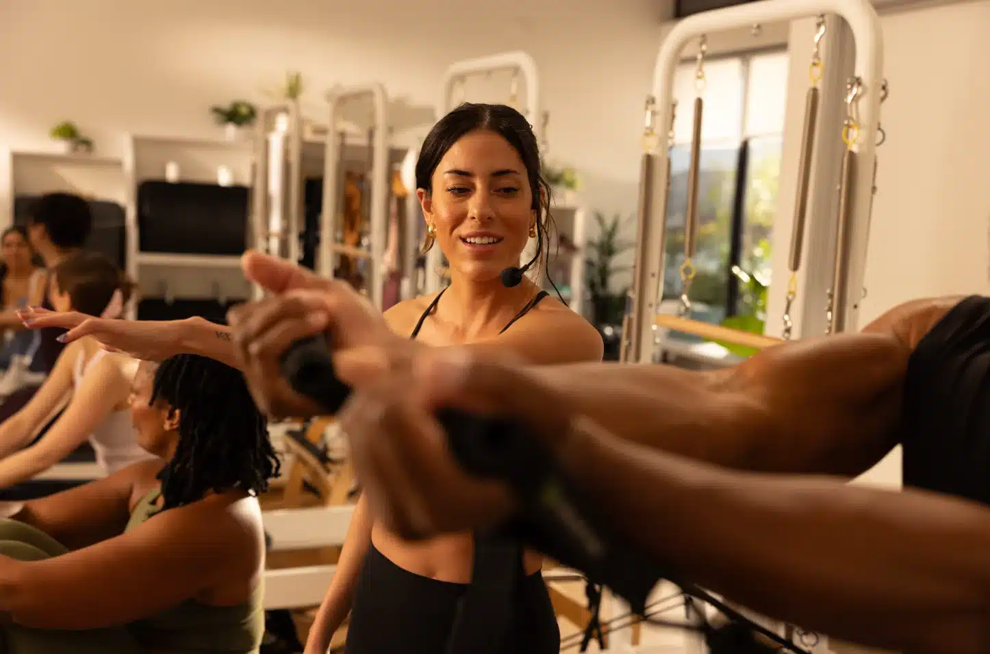 A woman instructs a Pilates class, smiling as participants use resistance bands in a well-lit studio.