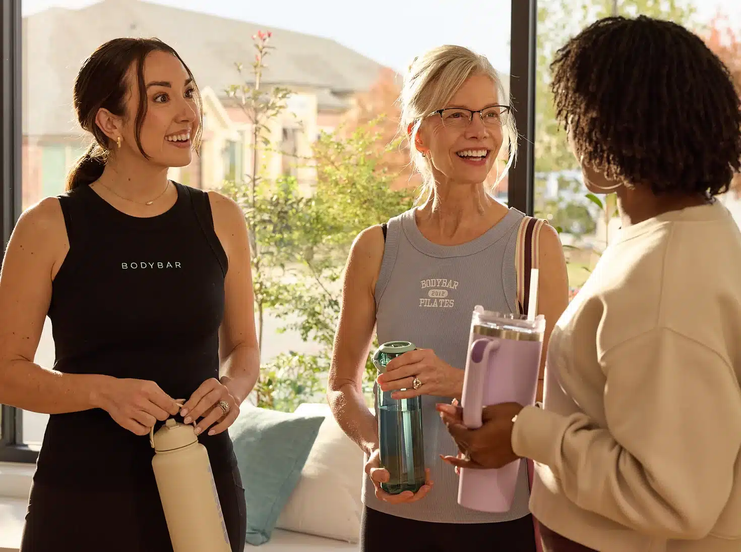 Three women in workout clothes stand indoors, smiling and talking while holding water bottles.