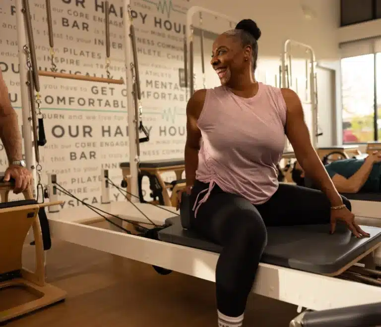 Smiling woman in workout clothes sits on Pilates reformer in a bright fitness studio with wall decor.