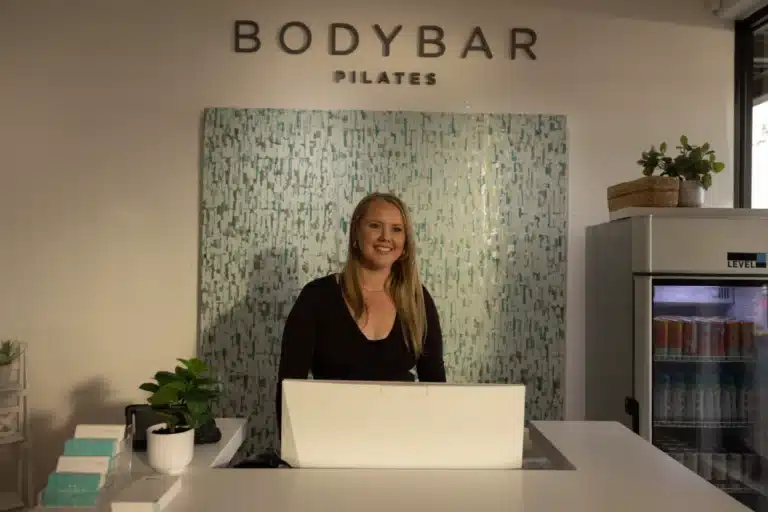Woman stands smiling behind a reception desk at a Pilates studio with plants and a fridge nearby.