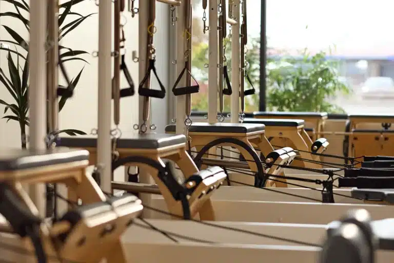 Pilates reformer machines lined up in a bright studio with large windows and green plants.