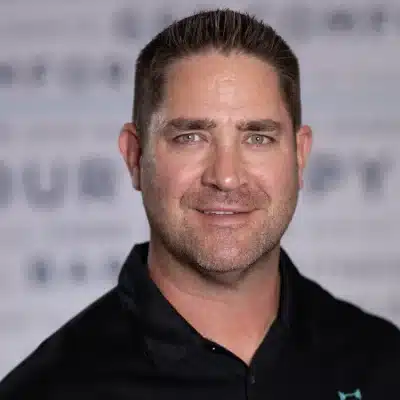 Man with short brown hair and trimmed beard wearing a black shirt, smiling at the camera with a blurred background.