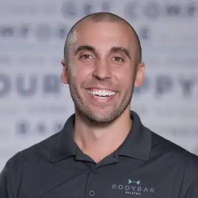 Smiling man with short hair wearing a dark BODYBAR Pilates shirt, standing in front of a blurred background.