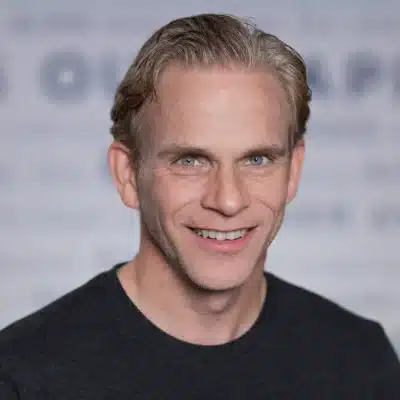 Smiling man with short light hair wearing a black shirt, in front of a blurred, light-colored background.