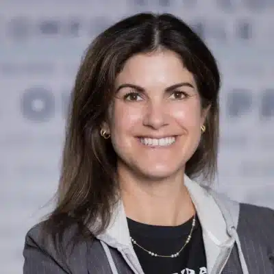 Woman with long brown hair, wearing a blazer and necklace, smiling in front of a blurred gray background.