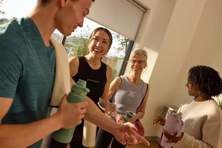 Four people in workout clothes smile and talk together, each holding a water bottle in a brightly lit room.