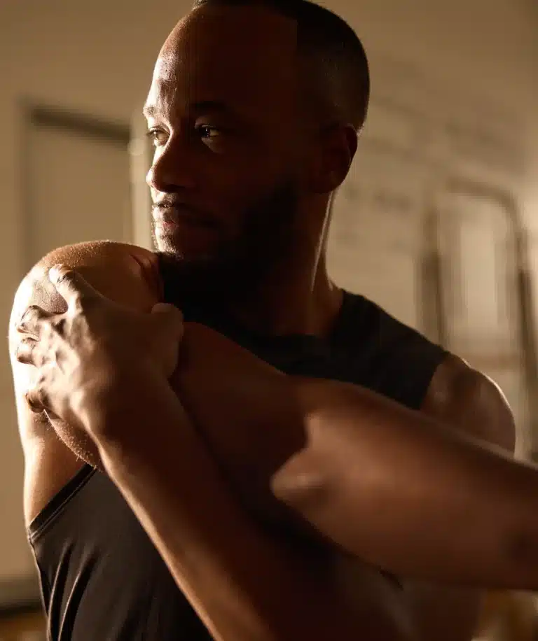Man in a black tank top stretching his arm across his chest, looking to the side in warm indoor lighting.
