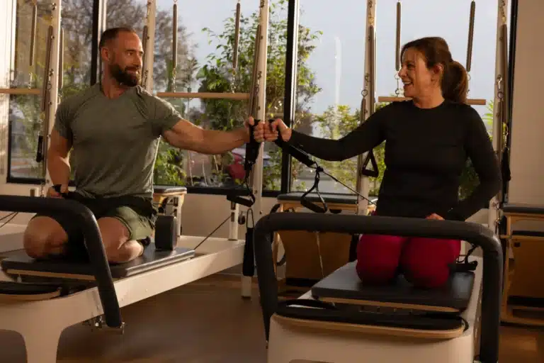 A man and woman kneel on Pilates reformers, smiling and high-fiving in a bright gym with large windows and plants.