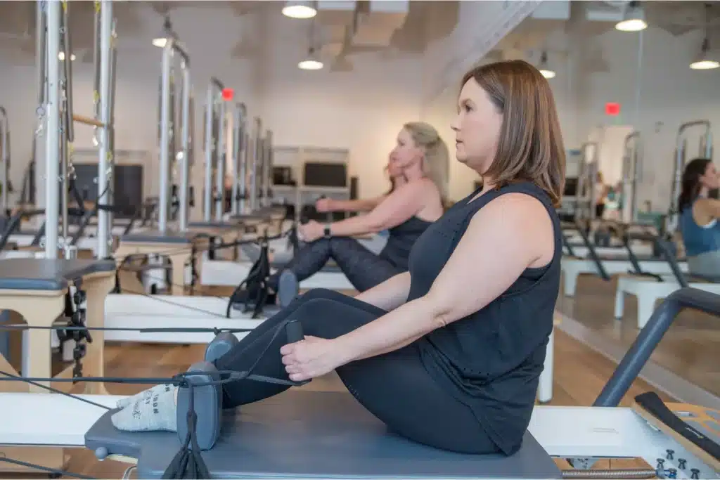Three women sit on Pilates reformers in a fitness studio, exercising and focusing on their workout.