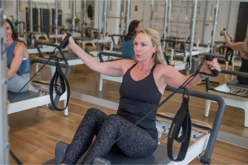 Woman in workout clothes using a Pilates reformer machine in a fitness studio, pulling on resistance straps.