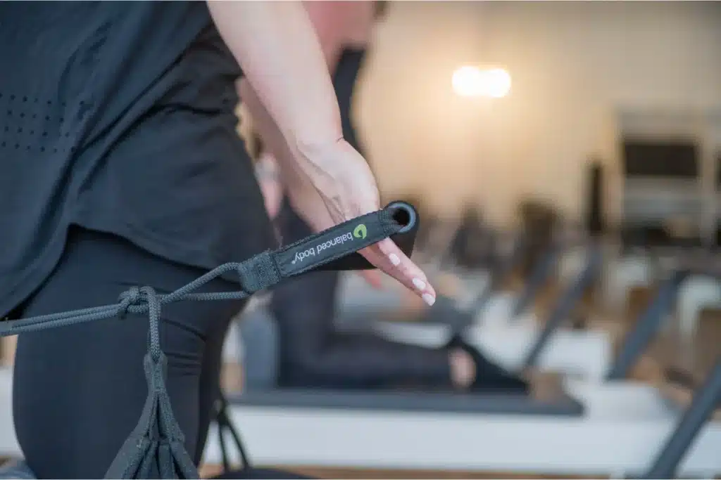 Close-up of a person holding a reformer Pilates strap in a fitness studio, with others exercising in the background.