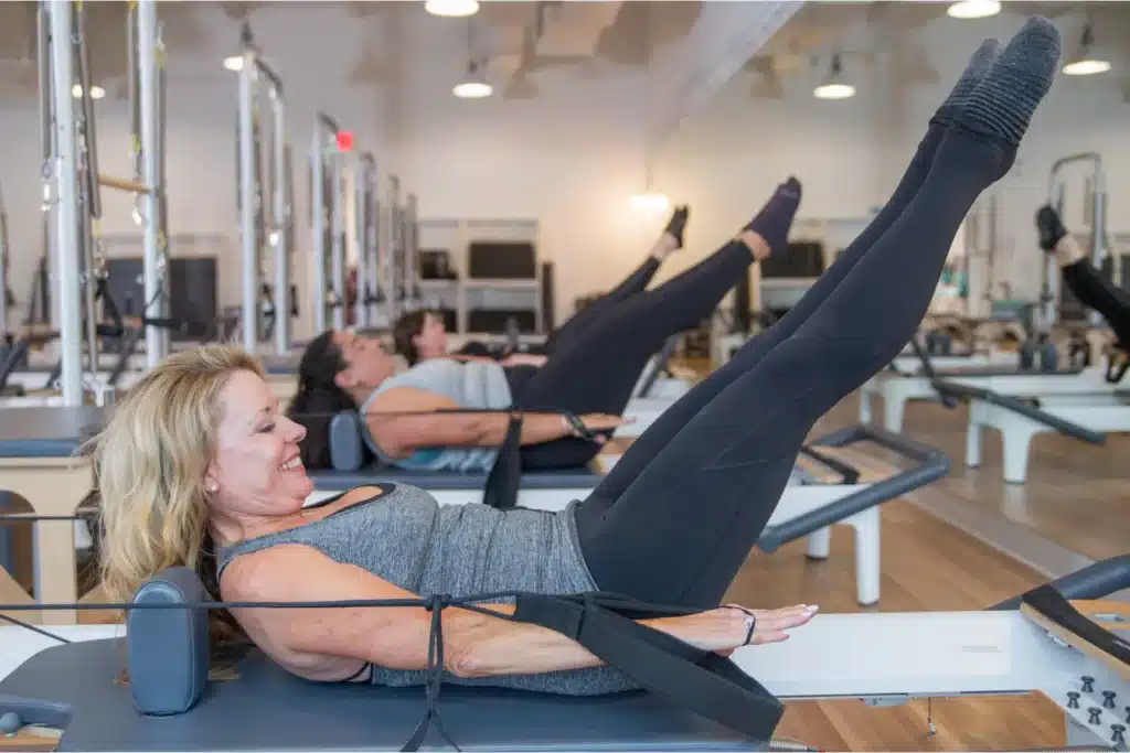 Women exercising on Pilates reformer machines, lying down with legs raised, in a bright fitness studio.