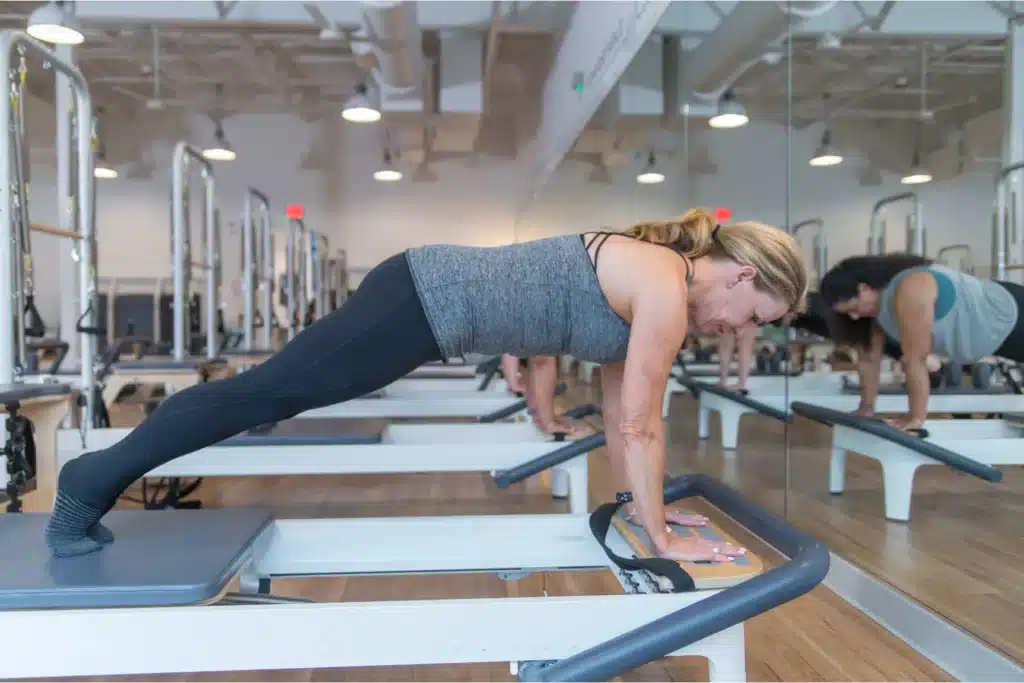 Woman doing a plank on a Pilates reformer in a fitness studio with mirrors and exercise equipment.