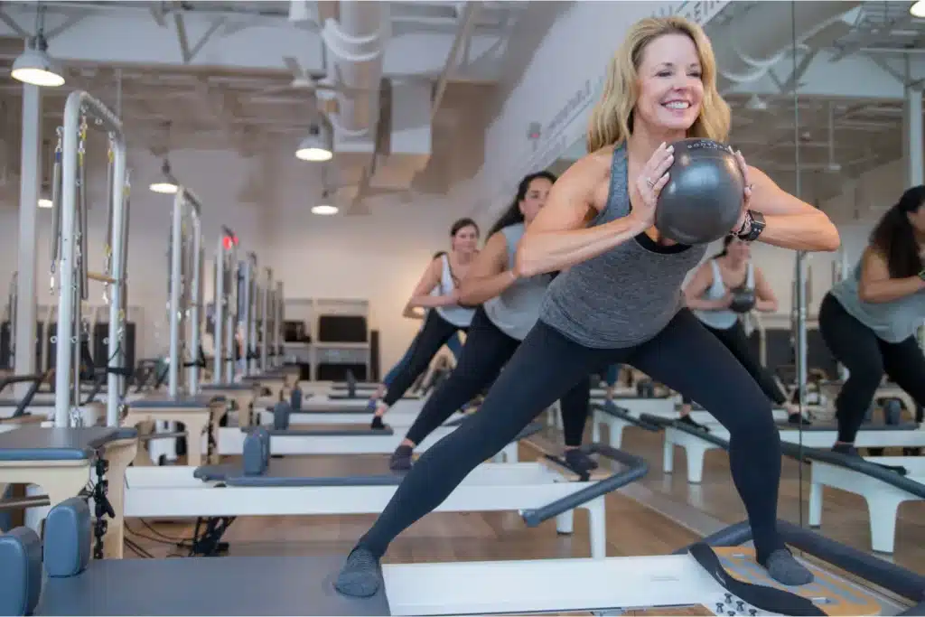 Women in workout clothes doing side lunges with medicine balls in a bright Pilates studio, smiling and focused.