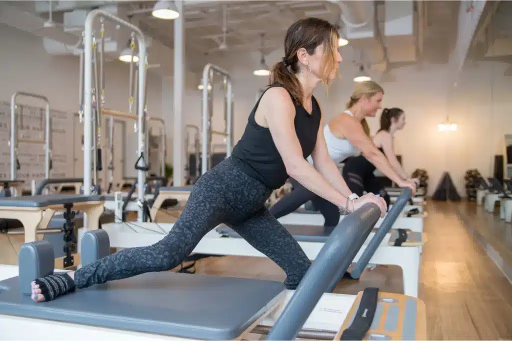 Three women exercise on Pilates reformer machines in a bright studio, stretching their legs and focusing ahead.