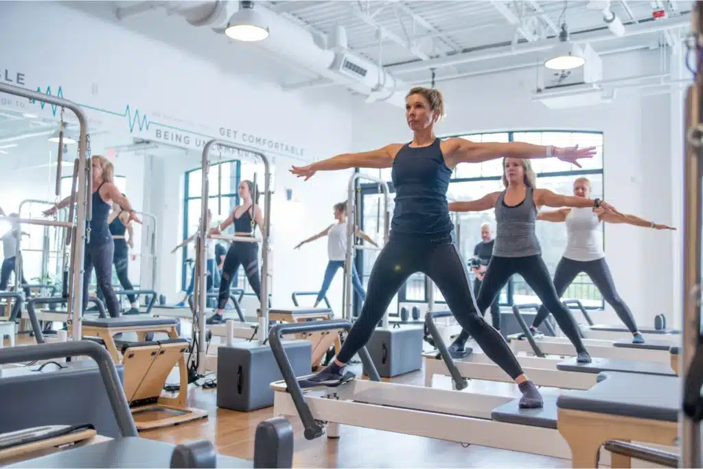 Women in workout clothes doing Pilates exercises on reformer machines in a bright, modern fitness studio.