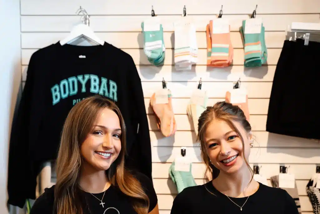 Two smiling women standing in front of a display wall with socks and a “BODYBAR” sweatshirt hanging on a hanger.