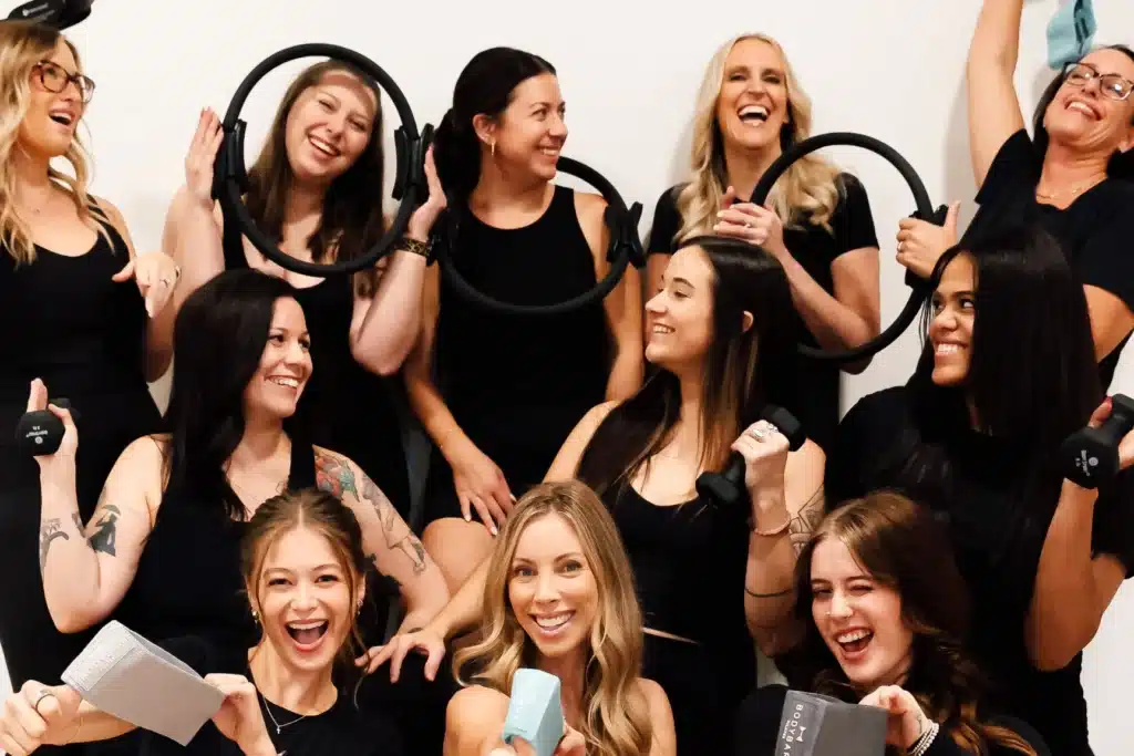 Nine women in black workout clothes smiling and laughing, holding exercise equipment in a fitness studio.