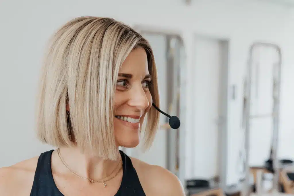 Woman with a blond bob haircut smiles while wearing a headset microphone in a bright indoor setting.