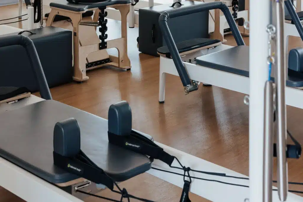 Several Pilates reformer machines are arranged in a studio with wooden floors and natural lighting.