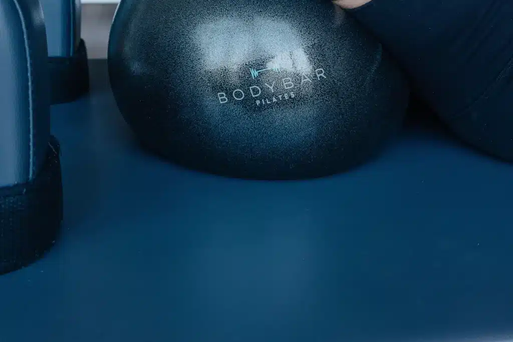 A black Pilates ball labeled “Body Bar” rests on a blue mat next to exercise equipment and an arm in a dark sleeve.