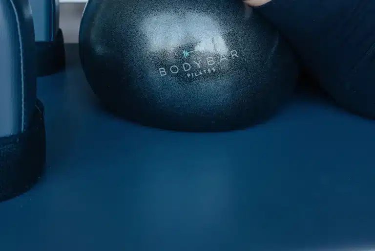 A black Pilates ball labeled “Body Bar” rests on a blue mat next to exercise equipment and an arm in a dark sleeve.