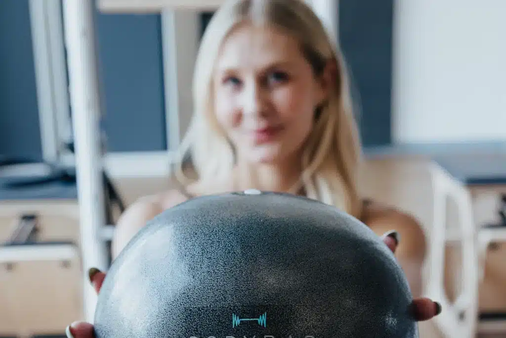 A woman holds a round gray medicine ball towards the camera in a gym setting, with her face out of focus.