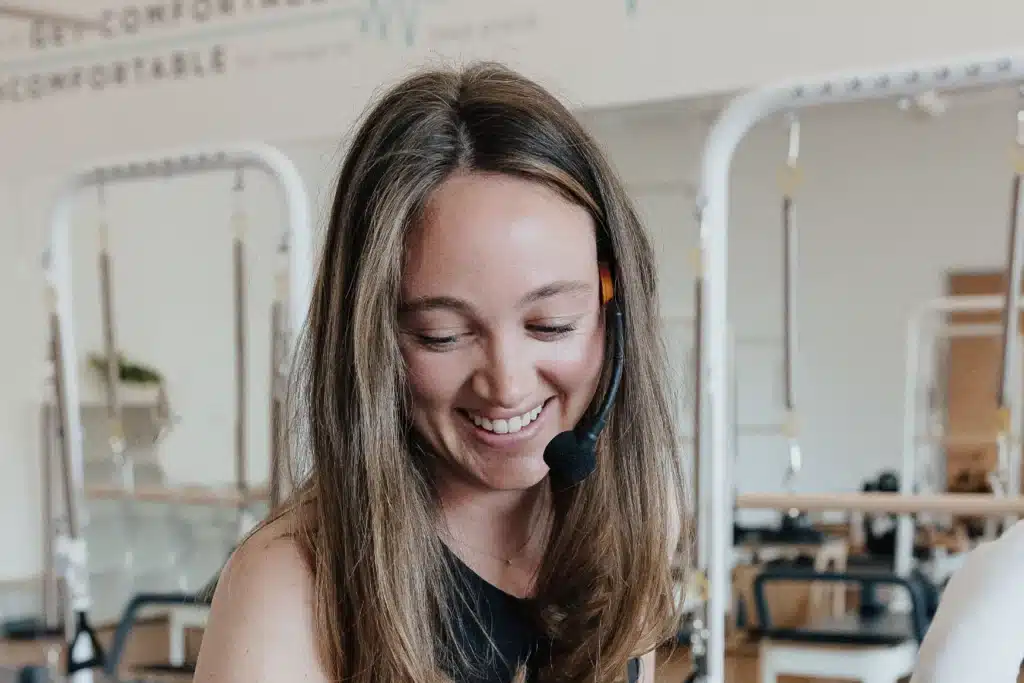 Woman with long brown hair, wearing a headset microphone, smiling in a fitness studio with pilates equipment in the background.
