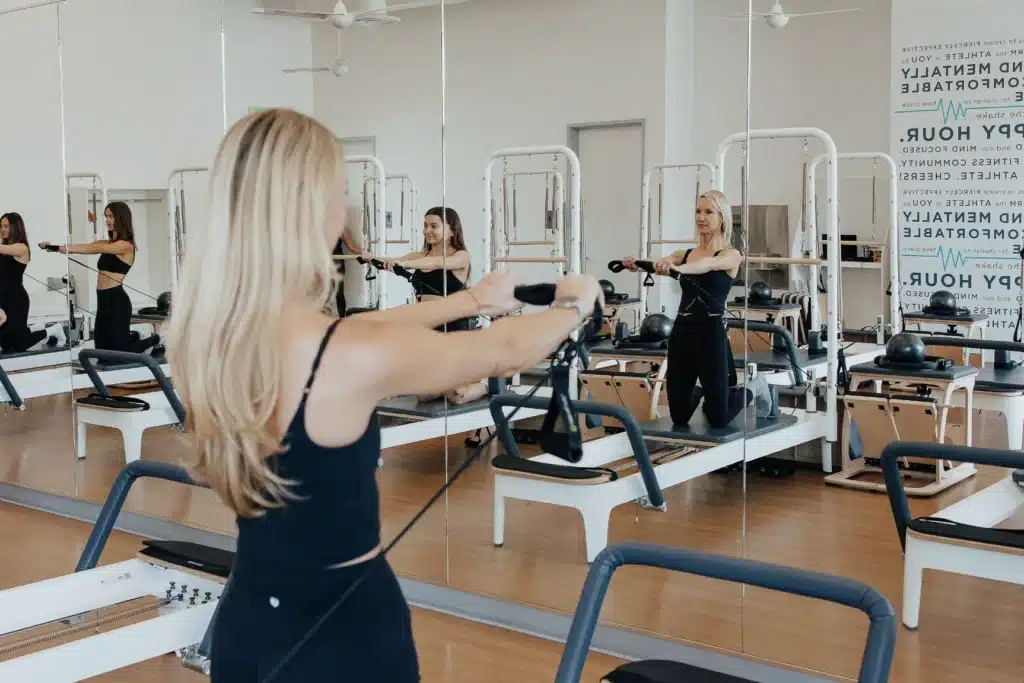 Woman exercising on a Pilates reformer in a fitness studio with mirrored walls and wooden floors.