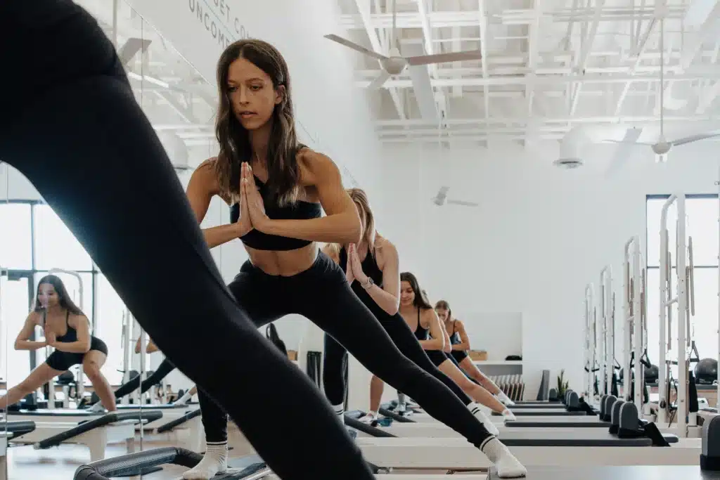 Women in athletic wear doing side lunges on reformer machines in a bright, modern fitness studio.