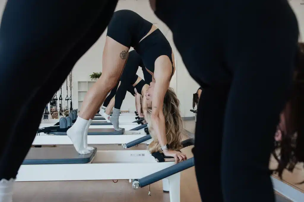 Women in athletic wear practice Pilates on reformer machines in a fitness studio, bending forward in a stretch.
