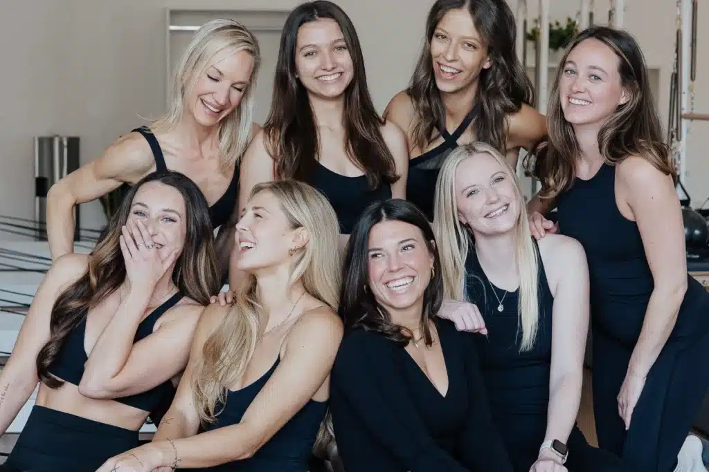 Eight women in black athletic wear sit and stand, smiling and laughing together in a bright indoor space.
