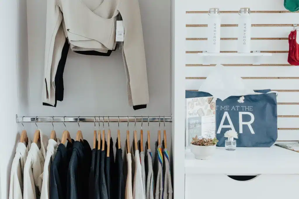 Clothes on wooden hangers, folded tops above, a blue tote bag, tissue box, and two bottles on a shelf nearby.