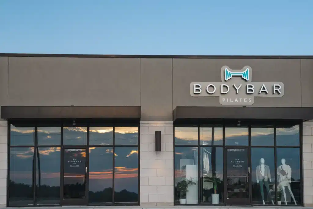 BODYBAR Pilates studio exterior at sunset, glass doors, mannequins, and fitness equipment visible inside.