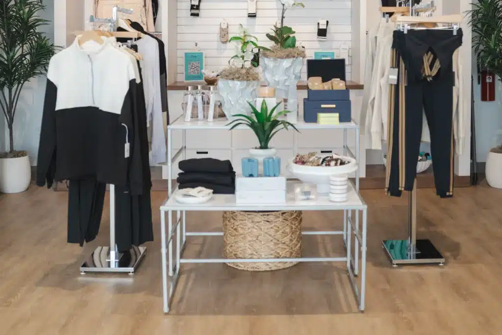 Clothing store display with black and white outfits, green plants, and neatly arranged accessories on tables.