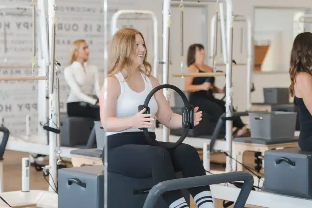 Woman in workout clothes uses a Pilates ring in a bright fitness studio with others exercising in the background.