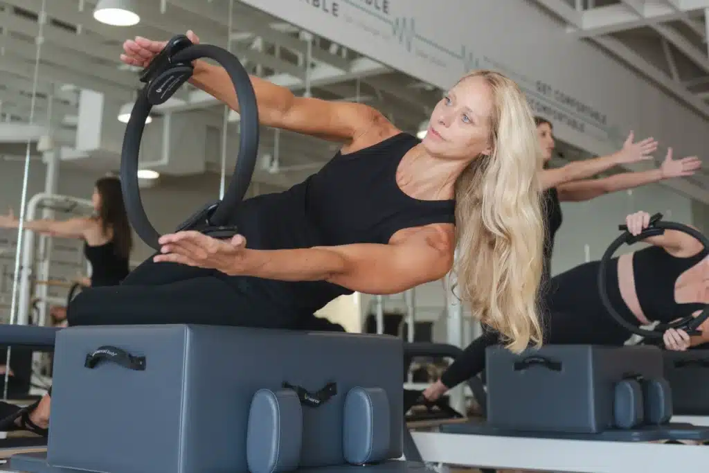 Women in a fitness studio use Pilates equipment, stretching with rings while seated on gray platforms.