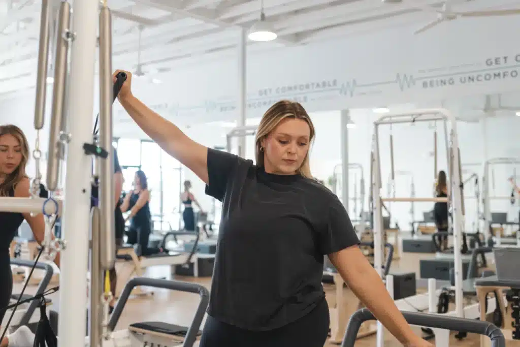 Woman in black shirt uses exercise equipment in a bright fitness studio with several people in the background.
