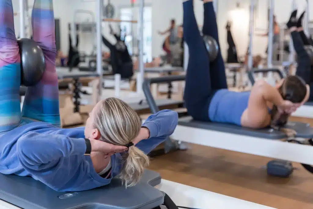Two women in athletic wear do Pilates exercises on reformers in a bright fitness studio with mirrors.